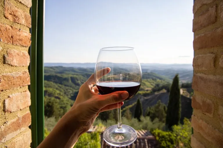 Frau hält ein Glas Rotwein mit schöner italienischer Landschaft im Hintergrund an einem sonnigen Tag. Blick aus dem Fenster.